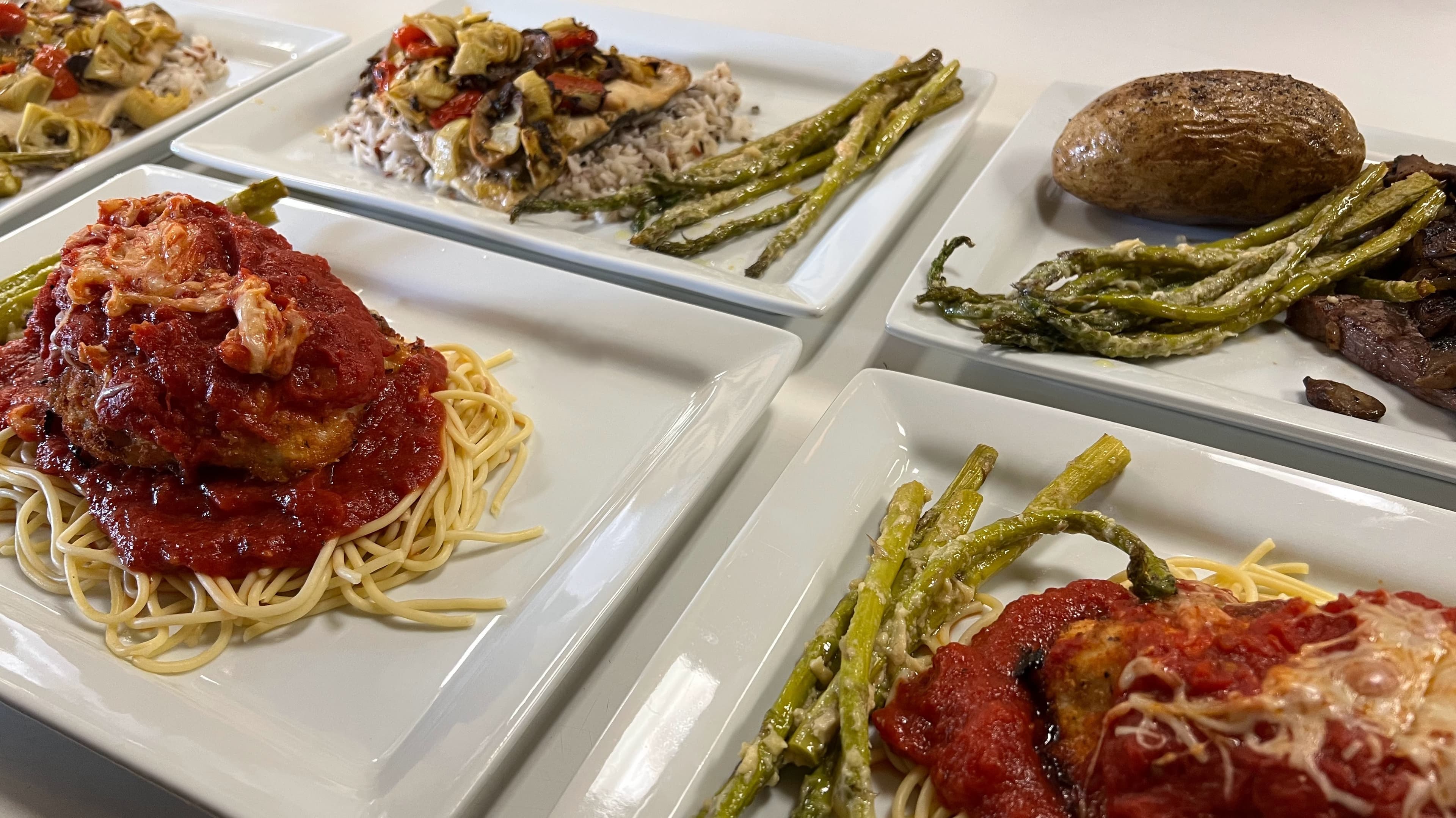 A selection of plated meals including spaghetti with marinara sauce, asparagus, a baked potato, and a dish of chicken with vegetables.