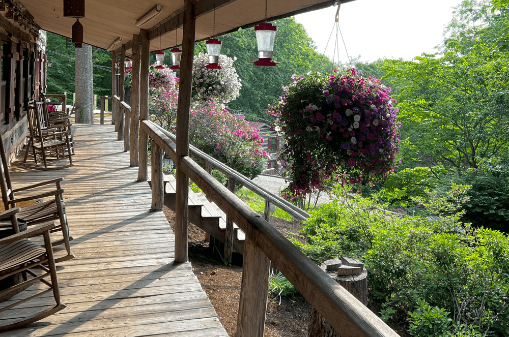 A wooden porch lined with rocking chairs and vibrant hanging flower baskets overlooks a lush green landscape.