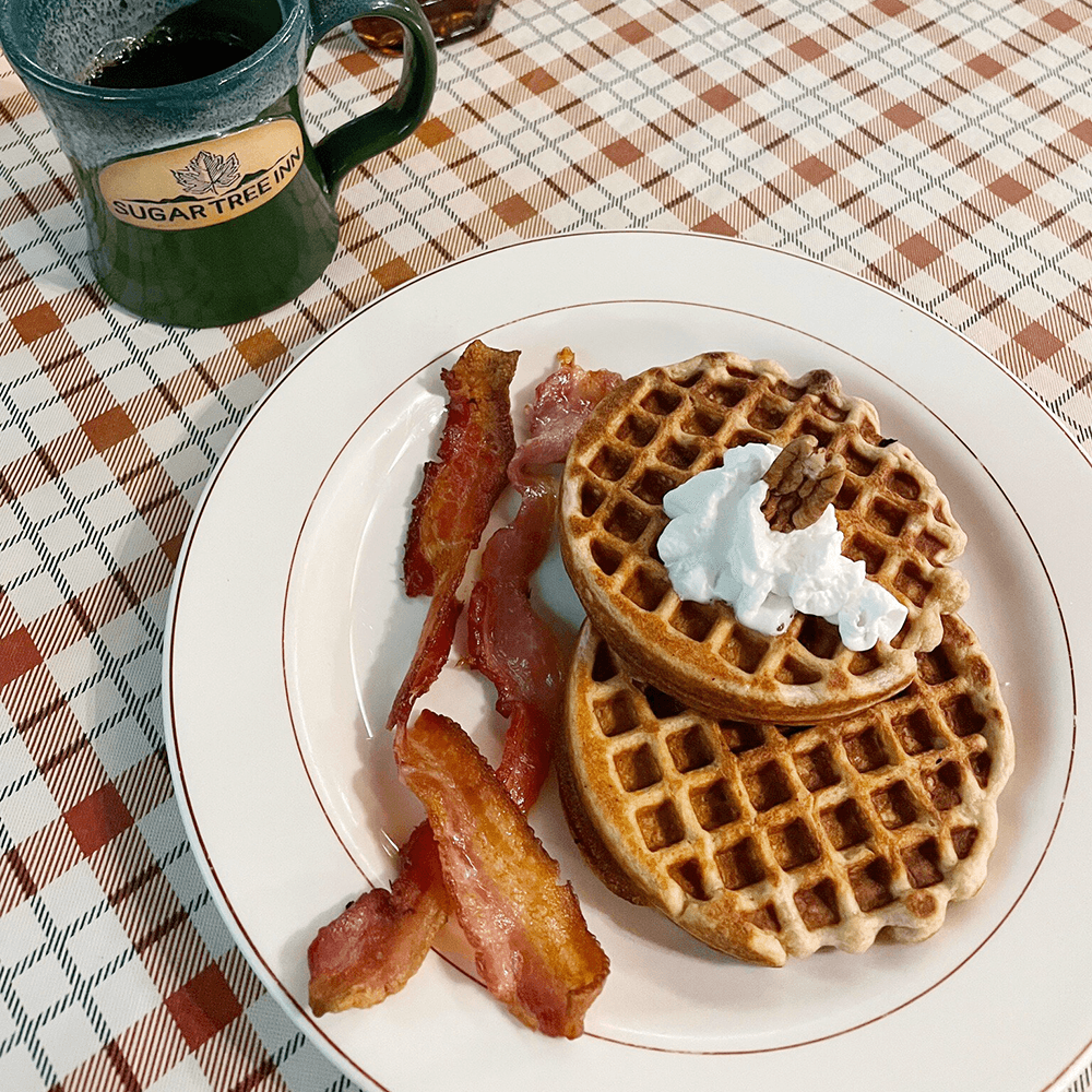 A plate of waffles topped with whipped cream and a pecan, accompanied by crispy bacon, beside a green mug of coffee.