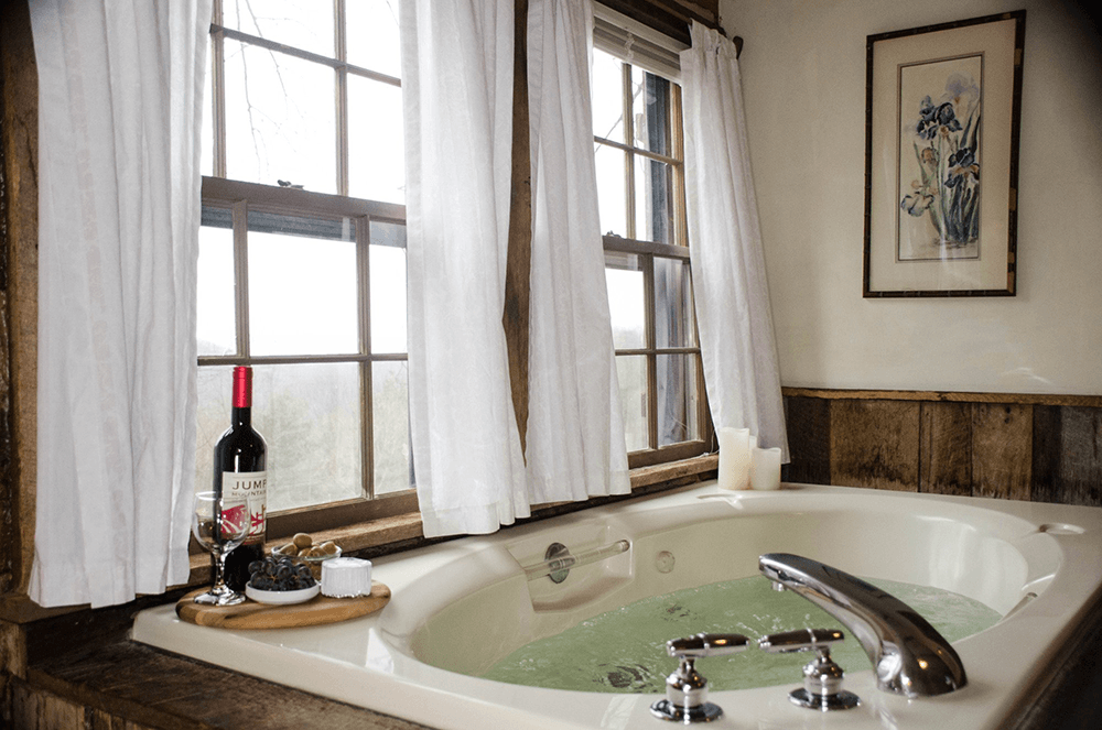 A cozy bathroom featuring a whirlpool tub, a bottle of red wine, glasses, and a scenic view from the windows.