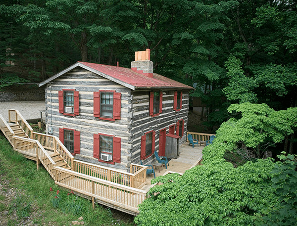 A two-story log cabin with red shutters surrounded by lush green trees and a wooden deck.
