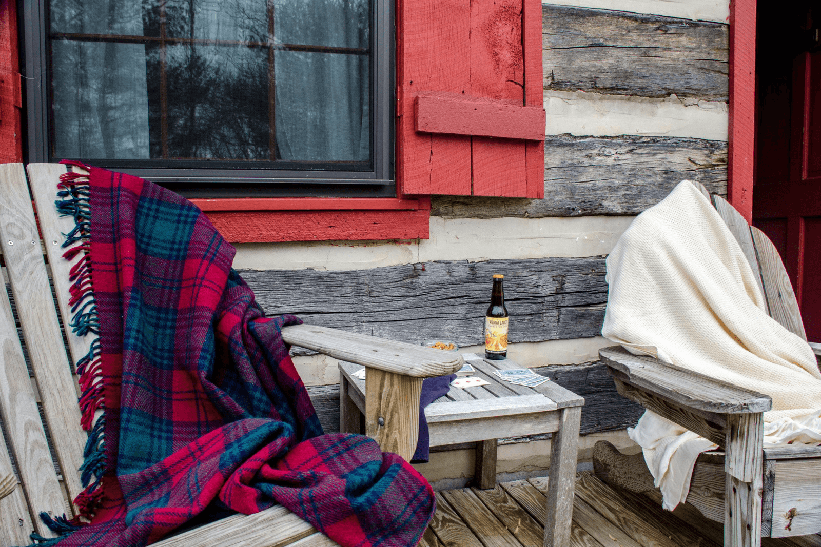 Two rustic chairs draped with blankets sit beside a small table holding a bottle and some snacks, against a log cabin backdrop.