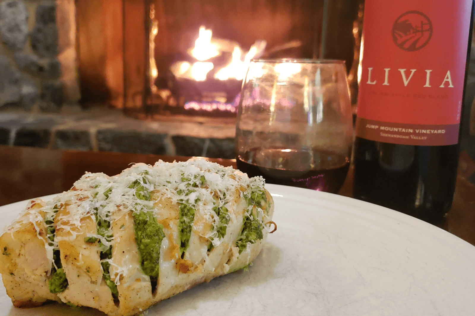 Spinach stuffed stromboli covered in shredded parmesan cheese on a plate, in front of a fire place, with a bottle of red wine and a half-full wine glass next to the plate